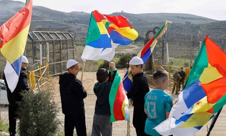 فتيان دروز يحملون أعلامًا بالقرب من جنود إسرائيليين يحرسون حاجزًا حدوديًا في قرية مجدل شمس بينما يُنتظر وصول وفد من الجانب السوري. 14 مارس 2025. ا ف ب
